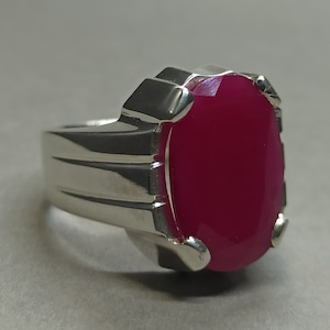 May include: A silver ring with a large, oval-shaped, bright pink gemstone set in a prong setting. The band of the ring has a textured design.