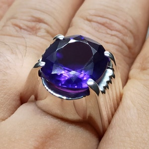 May include: A silver ring with a large, round, deep purple amethyst gemstone. The ring has a thick band with a textured design.