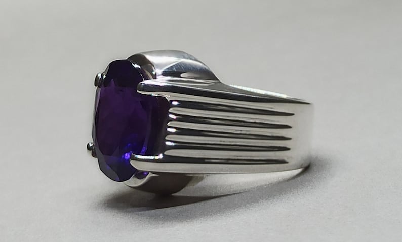 May include: A silver ring with a large, oval-shaped amethyst gemstone. The band of the ring has a textured, ridged design.