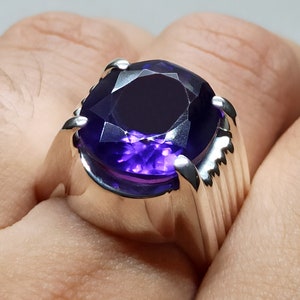 May include: A silver ring with a large, deep purple gemstone. The ring has a wide band with a textured design.