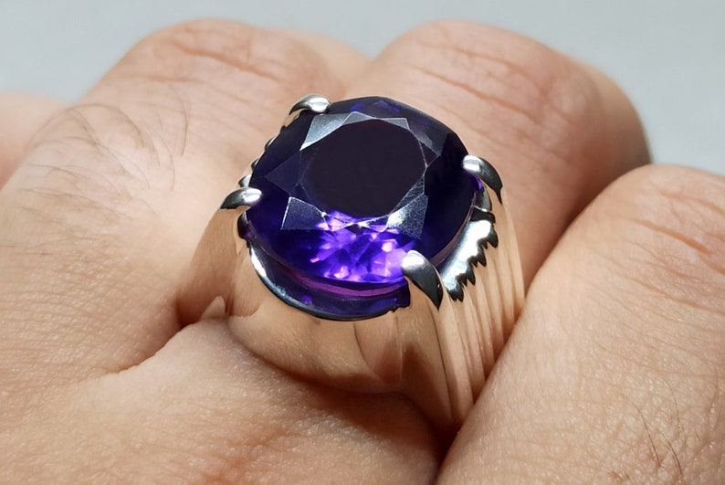 May include: A silver ring with a large, deep purple gemstone. The ring has a wide band with a textured design.