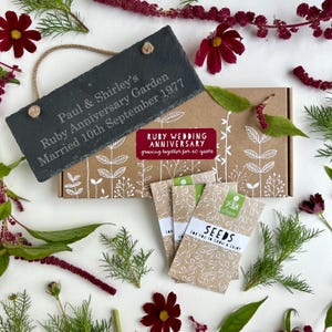 May include: A dark grey slate sign with engraved text, including a wedding anniversary date, sits atop a box with floral designs. Seed packets and red flowers are arranged around the box.