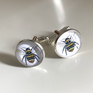 Könnte beinhalten: Ein Paar silberne Manschettenknöpfe mit weißem Hintergrund und einer Illustration einer schwarzen und gelben Biene.