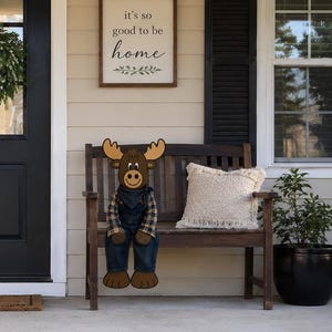 May include: A plush moose doll wearing a plaid shirt and denim overalls sits on a wooden bench on a porch. The porch features a black door with a wreath, a vertical wooden sign reading WELCOME, and a wall sign reading it's so good to be home.