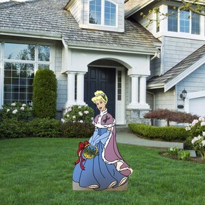 May include: A cardboard cutout of a princess in a blue gown and a pink fur-trimmed cape holding a basket of holly. The cutout is standing on a green lawn in front of a house.