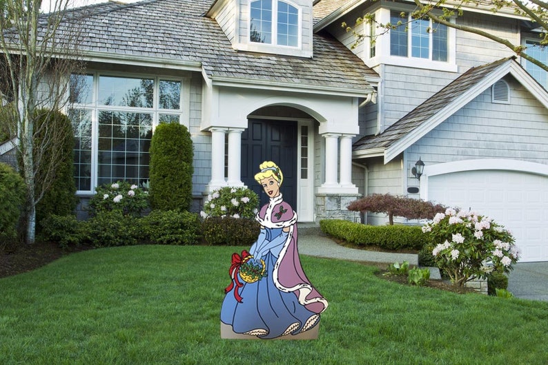 May include: A cardboard cutout of a princess in a blue gown and a pink fur-trimmed cape holding a basket of holly. The cutout is standing on a green lawn in front of a house.