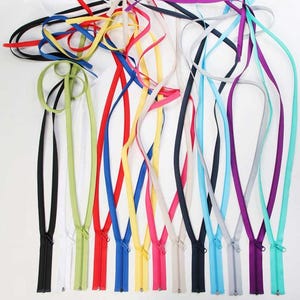 Puede incluir: Una colección de cremalleras coloridas en varios tonos, incluyendo negro, blanco, rojo, azul, amarillo, rosa y morado. Cada cremallera tiene una lengüeta de tela a juego. Las cremalleras están dispuestas sobre un fondo blanco.