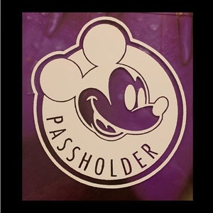 May include: A white and purple circle with a cartoon image of Mickey Mouse's head and the word "PASSHOLDER" written in a white circle around the image.