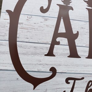 May include: Close-up of a brown wooden sign with the word "Camp" in a decorative font. The sign has a whitewashed background and the letters are raised.