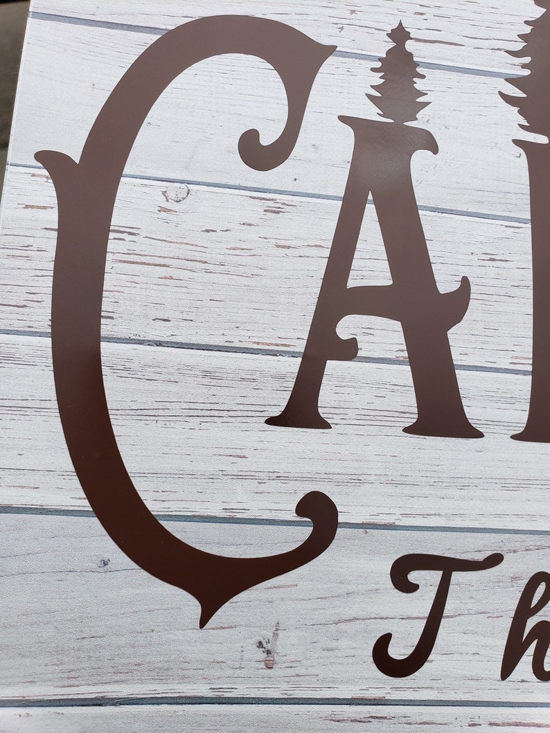 May include: Close-up of a brown wooden sign with the word "Camp" in a decorative font. The sign has a whitewashed background and the letters are raised.