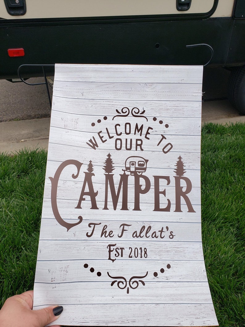 May include: White wooden sign with brown text that reads "Welcome to our Camper The Fallat's Est 2018". The sign has a stylized camper icon and a decorative flourish.