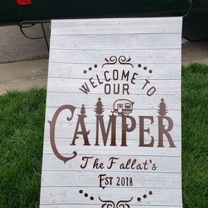 May include: White wooden sign with brown text that reads "Welcome to our Camper The Fallat's Est 2018". The sign has a brown camper silhouette and pine trees.