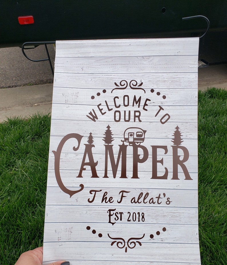 May include: White wooden sign with brown text that reads "Welcome to our Camper The Fallat's Est 2018". The sign has a brown camper silhouette and pine trees.