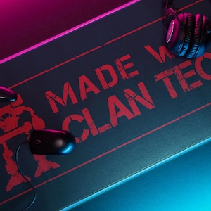 May include: Gaming setup with a black mouse, controller, and headphones on a black mousepad. The mousepad has red text reading "MADE WITH CLAN TECH" and a red robot graphic. The setup is lit with blue and pink lights.