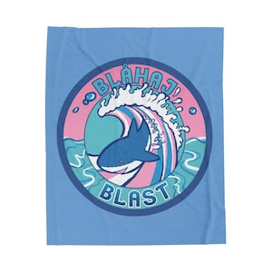 Könnte beinhalten: Eine blaue Decke mit einem kreisförmigen Design, das einen Hai zeigt, der eine Welle reitet. Das Design enthält den Text "BLÅHAJ" über der Welle und "BLAST" darunter, vor einem rosa-blauen Hintergrund.