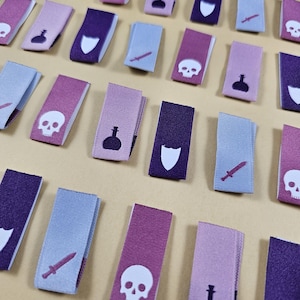 May include: A collection of purple, pink, and blue fabric labels with various symbols, including a skull, a sword, a potion bottle, and a shield. The labels are arranged in rows on a beige surface.