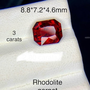 May include: A faceted, rectangular red rhodolite garnet gemstone. The text on the image reads 8.8*7.2*4.6mm, 3 carats, and Rhodolite garnet. The stone displays deep red hues with sharp, geometric facets that reflect light across its surface.
