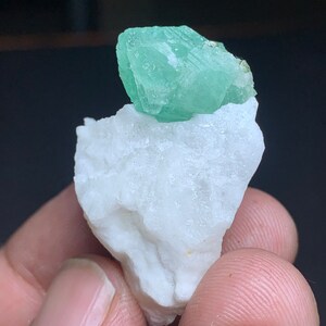 Op de afbeelding: Een klein, groen fluorietkristal op een witte steen. Het kristal is doorschijnend en heeft een glazige glans.