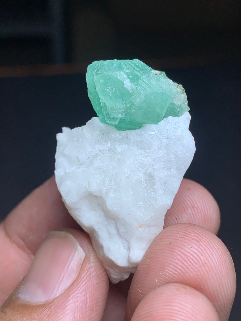 Op de afbeelding: Een klein, groen fluorietkristal op een witte steen. Het kristal is doorschijnend en heeft een glazige glans.