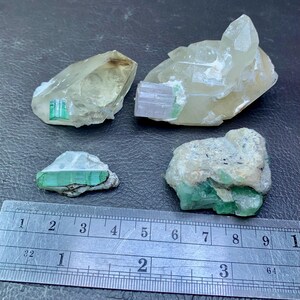 Może przedstawiać: Cztery surowe okazy minerałów, w tym kwarc i turmalin, są prezentowane na ciemnej powierzchni. Kryształy różnią się rozmiarem i kolorem, z zielonymi i jasnymi odcieniami. Obok minerałów umieszczono linijkę dla skali.