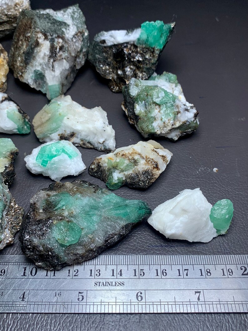 Op de afbeelding: Een verzameling ruwe smaragd kristallen in verschillende maten, vari&euml;rend van klein tot groot, ingebed in een witte en grijze matrix. De kristallen zijn een levendige groene kleur en hebben een glazige glans. Een liniaal is inbegrepen voor de schaal.
