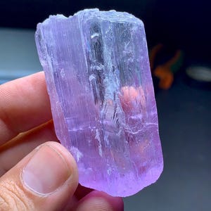 May include: A translucent, lavender-colored crystal with a rectangular shape. The crystal has a rough, textured surface and appears to be held by a person. The crystal's color ranges from pale to deep purple, with some areas appearing almost clear.