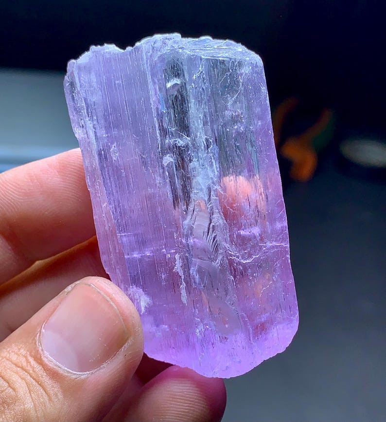 May include: A translucent, lavender-colored crystal with a rectangular shape. The crystal has a rough, textured surface and appears to be held by a person. The crystal's color ranges from pale to deep purple, with some areas appearing almost clear.