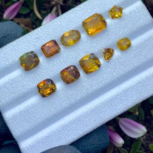 Może przedstawiać: Kolekcja fasetowanych kamieni szlachetnych o różnych kształtach, w tym owalnych, kwadratowych i prostokątnych. Kamienie prezentują ciepłe kolory, od złotożółtego po bursztynowy i brązowy. Ułożone są na białej powierzchni.