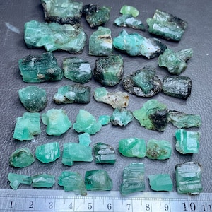 Op de afbeelding: Een verzameling van 30 ruwe smaragd kristallen in verschillende maten en vormen. De kristallen zijn een levendige groene kleur en hebben een natuurlijke, onbewerkte afwerking. De kristallen zijn gerangschikt op een grijs oppervlak met een liniaal voor schaal.
