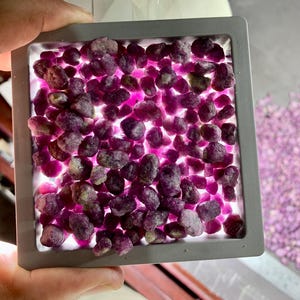 Può includere: Una collezione di gemme grezze, principalmente nelle tonalità del viola e del rosa, è esposta all'interno di un contenitore quadrato con cornice grigia. Le pietre sono illuminate dal basso, creando un effetto luminoso. L'immagine è tenuta da una mano.