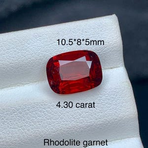 May include: A cushion-cut, deep red rhodolite garnet gemstone with a faceted surface. The stone is labeled with the text 10.5*8*5mm, 4.30 carat, and Rhodolite garnet. The gemstone displays a vibrant, saturated red color and a polished finish.