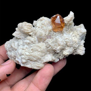 Może przedstawiać: Naturalny okaz minerału z skupiskiem białych, teksturowanych formacji skalnych. Centralnie umieszczony, fasetowany, bursztynowy kryształ, z widocznymi mniejszymi kryształami. Tło jest jednolicie czarne, podkreślające szczegóły minerału.