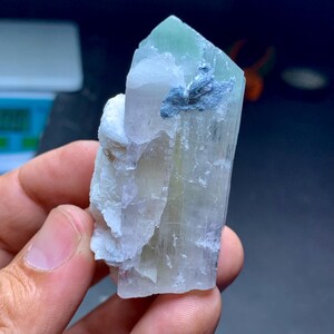 May include: A translucent crystal formation with a pale green tip and a light blue inclusion. The crystal has a layered appearance and is held in a hand. The overall color palette is soft and natural.