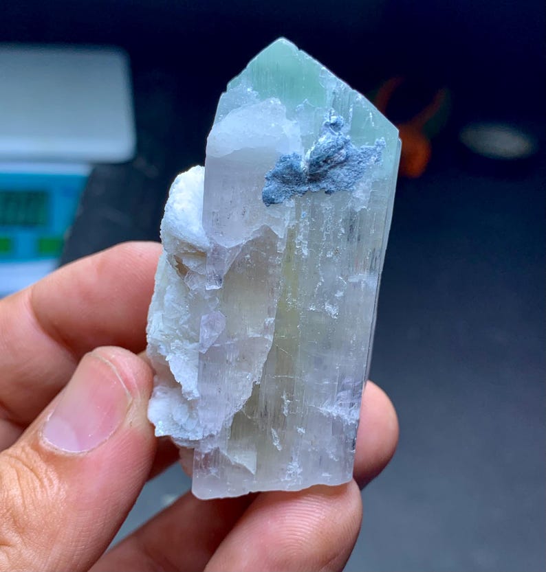 May include: A translucent crystal formation with a pale green tip and a light blue inclusion. The crystal has a layered appearance and is held in a hand. The overall color palette is soft and natural.