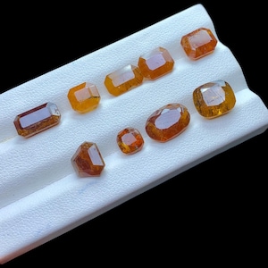 Könnte beinhalten: Eine Sammlung facettierter, orange-brauner Edelsteine in verschiedenen Formen und Größen. Die Edelsteine werden auf einer weißen, strukturierten Oberfläche vor schwarzem Hintergrund präsentiert. Die Edelsteine haben ein poliertes Aussehen und reflektieren Licht.