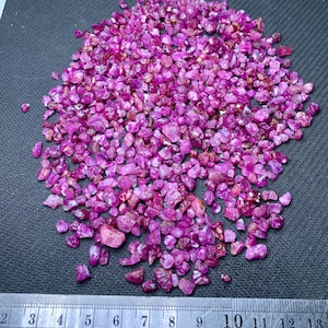 May include: A pile of raw, uncut ruby gemstones in varying shades of pink and red. The rough stones are scattered on a dark gray surface, with a metal ruler providing scale. The gemstones have a natural, unpolished appearance.