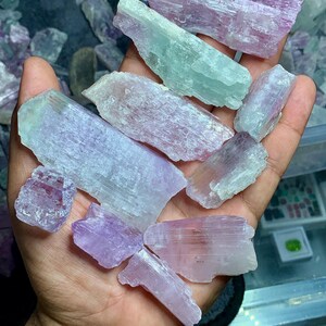 以下が含まれることがあります： ラベンダー、ピンク、ペールグリーンの原石の宝石の束。結晶はざらざらとした質感で、サイズも様々です。宝石は開いた手に持たれており、自然な美しさと色のバリエーションを示しています。