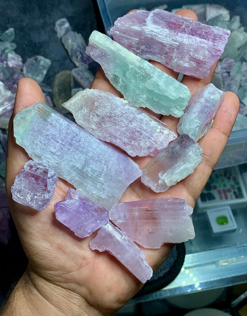 以下が含まれることがあります： ラベンダー、ピンク、ペールグリーンの原石の宝石の束。結晶はざらざらとした質感で、サイズも様々です。宝石は開いた手に持たれており、自然な美しさと色のバリエーションを示しています。