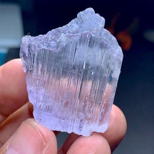 May include: A light purple, raw crystal with a textured surface. The crystal has vertical striations and a jagged top edge. The color is a pale lavender, and the overall shape is irregular. The crystal is held in a person's hand.