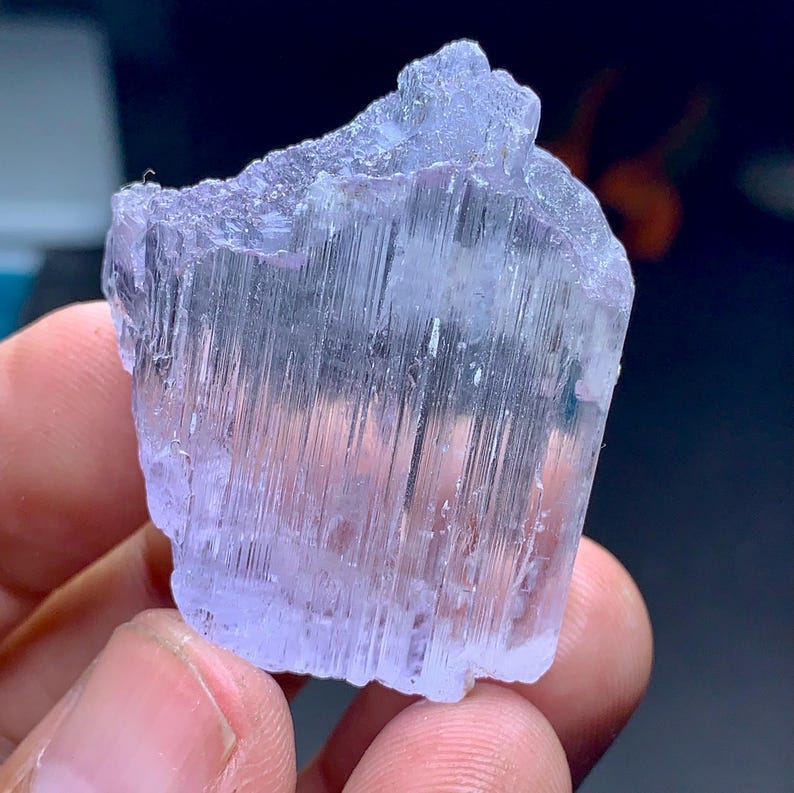 May include: A light purple, raw crystal with a textured surface. The crystal has vertical striations and a jagged top edge. The color is a pale lavender, and the overall shape is irregular. The crystal is held in a person's hand.