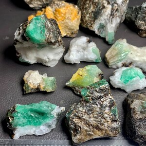 Op de afbeelding: Een verzameling ruwe smaragd kristallen in verschillende maten en vormen. De kristallen zijn een levendige groene kleur en zijn ingebed in een matrix van grijs en wit gesteente. Een liniaal is inbegrepen voor de schaal.