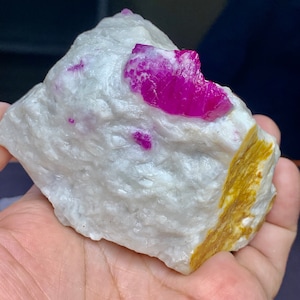 May include: A rough, white rock with a large, bright pink ruby crystal embedded in it. The rock has a yellow-brown section on one side.
