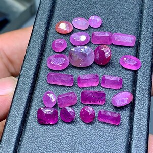 Op de afbeelding: Een assortiment gefacetteerde, levendige roze en rode edelstenen. De edelstenen variëren in vorm, waaronder ovaal, rond en rechthoekig. De stenen worden getoond op een donker, getextureerd oppervlak, dat hun glans en kleur benadrukt.