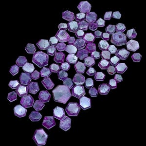 May include: A collection of numerous loose, flat, hexagonal-shaped purple and pink gemstones. Each stone features a translucent, polished surface with visible internal inclusions and varying shades of magenta, violet, and light pink hues.