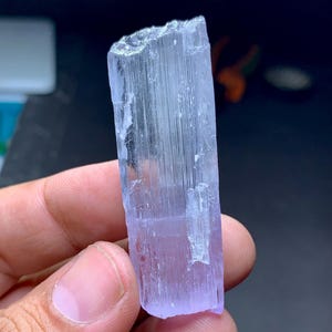 May include: A translucent, light purple crystal with a rectangular shape. The crystal has a rough, uneven top and a smooth, polished surface. The color transitions from a pale lavender to a clear, icy hue. The crystal is held in a hand.