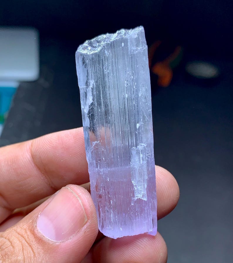 May include: A translucent, light purple crystal with a rectangular shape. The crystal has a rough, uneven top and a smooth, polished surface. The color transitions from a pale lavender to a clear, icy hue. The crystal is held in a hand.