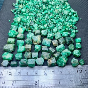 Può includere: Una vasta collezione di pietre preziose smeraldo grezze e non lucidate di varie forme e dimensioni, disposte sopra un righello metallico che misura fino a 10 centimetri, mostrando la scala delle pietre rispetto alle marcature metriche.