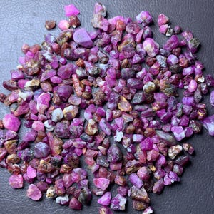 Puede incluir: Un montón grande de piedras brutas de rubí rosa y rojo. Las piedras brutas son de varios tamaños y formas. Una regla está en la parte inferior de la imagen para la escala.