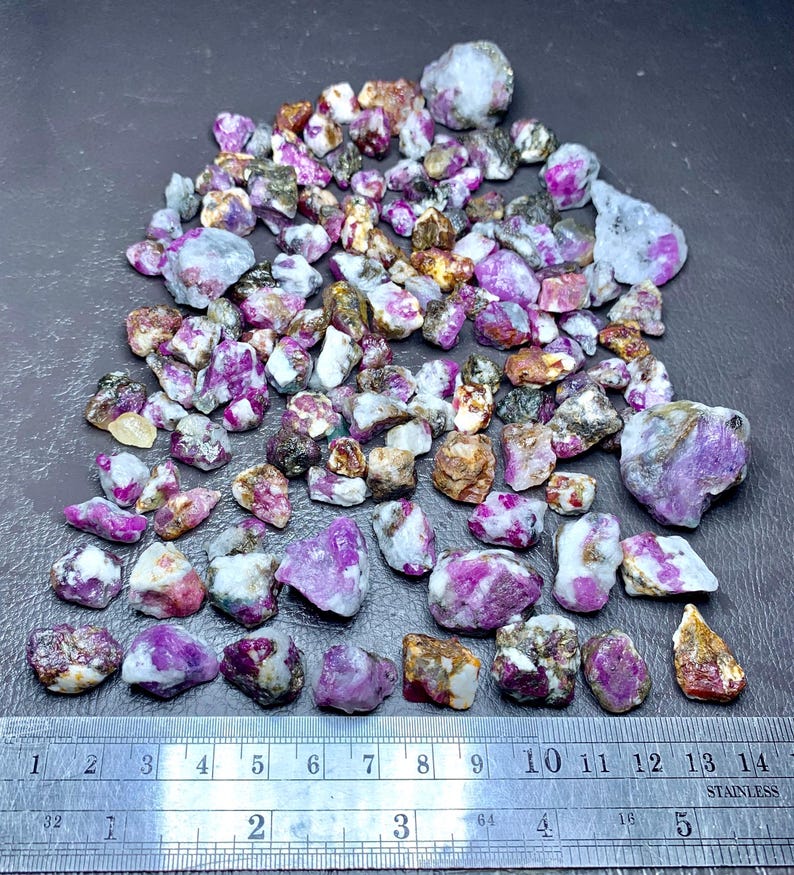 May include: A collection of raw, uncut ruby gemstones in various sizes and shapes. The stones display a range of colors, including deep pink, purple, and white, with some having brown and gray inclusions. A metal ruler with inch markings is at the bottom.