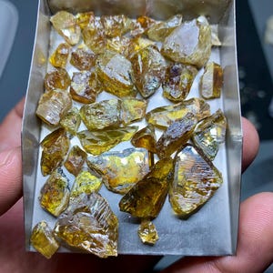 Op de afbeelding: Een verzameling ruwe, ongeslepen edelstenen in een metalen bakje. De edelstenen variëren in grootte en vorm en vertonen tinten geel, amber en bruin. De ruwe textuur en natuurlijke facetten van de stenen zijn zichtbaar.